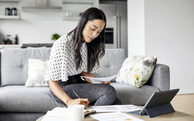 Young woman analyzing bills while writing in diary. Beautiful female is using digital tablet at table. She is sitting on sofa at home.